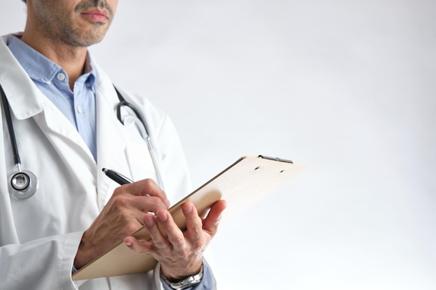 Jeune médecin méconnaissable avec blouse blanche et chemise bleue en semi-profil écrit sur un dossier avec fond blanc