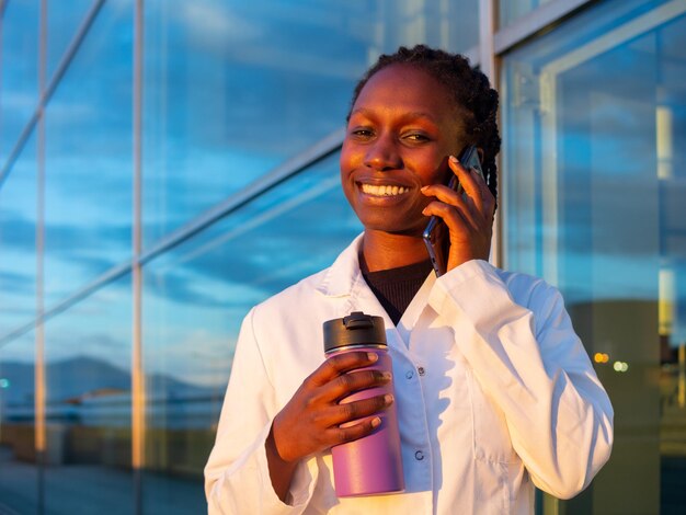 Un jeune médecin parle au téléphone à l'extérieur devant un hôpital en regardant la caméra.