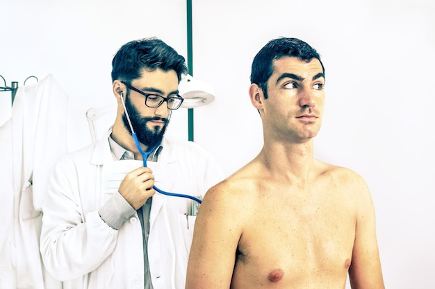 Photo jeune médecin primaire hipster au travail auscultant un patient masculin dans une clinique privée