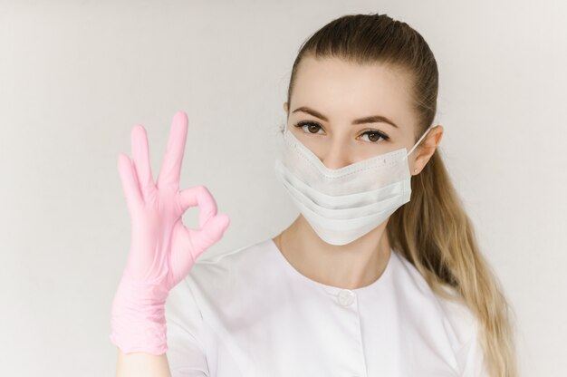 Jeune médecin prospère en masque de protection et blouse blanche montre signe avec une main correcte