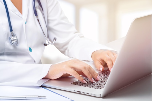 Jeune médecin sérieux en uniforme médical blanc assis au bureau à l'hôpital avec un ordinateur portable