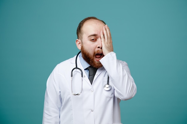 Jeune Médecin De Sexe Masculin Endolori Portant Un Manteau Médical Et Un Stéthoscope Autour Du Cou En Gardant La Main Sur Les Yeux En Regardant La Caméra Isolée Sur Fond Bleu