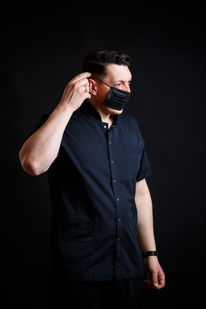 Un jeune médecin de sexe masculin vêtu d'une combinaison chirurgicale noire met un masque noir pour se protéger du virus. Isolé sur fond noir