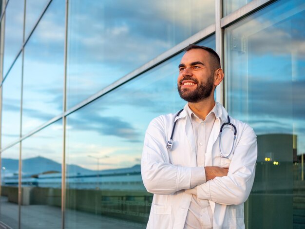 Un jeune médecin sourit et croise les bras à l'extérieur près d'un hôpital.