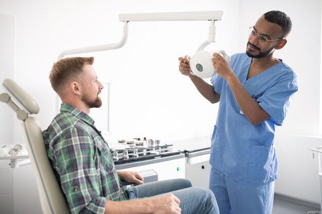 Jeune médecin en uniforme tenant un équipement médical ou une lampe en se tenant debout devant un patient dans un fauteuil avant l'examen