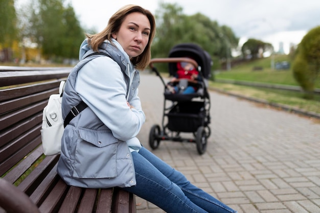 Jeune mère agacée assise dans le parc sur un banc loin de la poussette avec un visage en colère