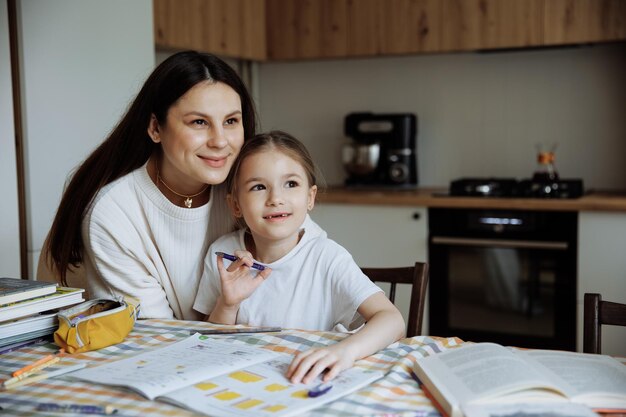 Une jeune mère aide sa fille de première année à faire ses devoirs à la maison dans la cuisine.