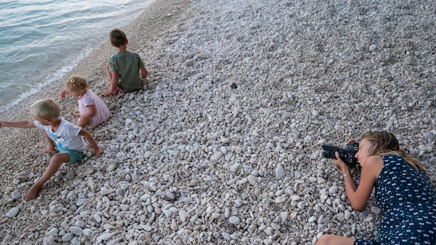 Jeune mère allongée sur une plage de galets le soir pour prendre des photos de ses trois enfants