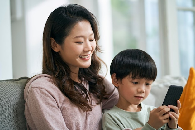 Une jeune mère asiatique assise sur le canapé de la famille à la maison partageant des images de téléphone portable avec son fils