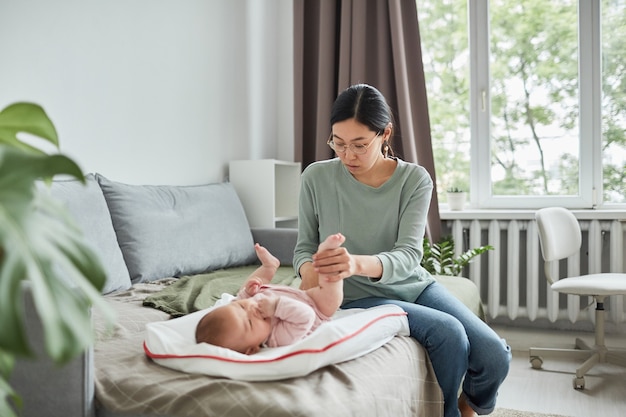 Jeune mère asiatique assise sur un canapé et s'occupant de son bébé pendant qu'elle est allongée sur le lit dans la chambre