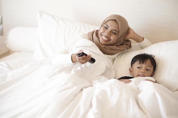 Jeune mère asiatique et son fils jouent joyeusement sur le lit tout en regardant la télévision dans la chambre d'hôtel en vacances