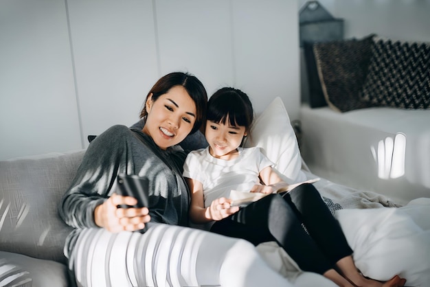 Une jeune mère asiatique utilise son smartphone et lit des livres à sa petite fille au lit. Ils apprécient le temps de lien familial à la maison.