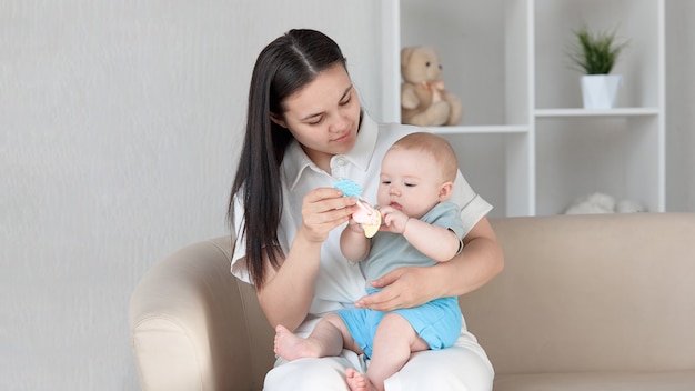 Une jeune mère, assise sur le canapé avec un bébé sur ses genoux, jouant avec son hochet pour bébé