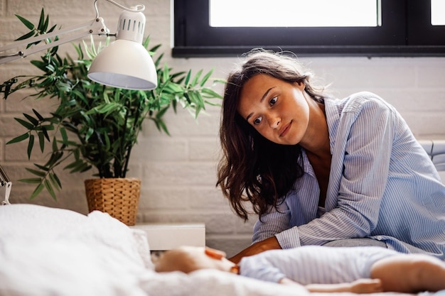 Photo jeune mère assise près du lit et regardant son bébé endormi