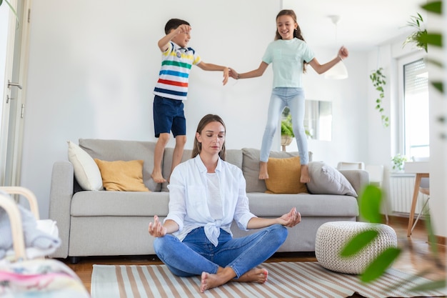 Jeune mère aux yeux fermés méditant dans une pose de lotus sur le sol en essayant de sauver l'harmonie intérieure tandis que des enfants excités sautent sur un canapé et crient dans un salon spacieux et lumineux