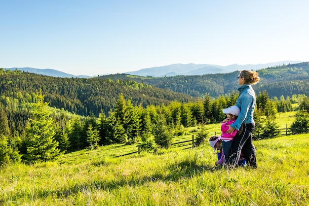 Jeune mère et deux petites filles voyageurs se tiennent sur une pente avec une vue magnifique sur les collines couvertes de forêt dense de sapins contre le ciel bleu