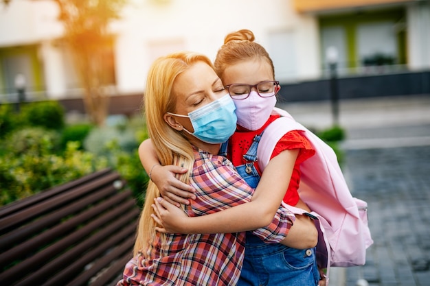 Jeune mère disant au revoir dans une cour d'école à sa petite fille avec un masque de protection faciale. Ils portent des masques de protection du visage. Retour au concept de l'école.