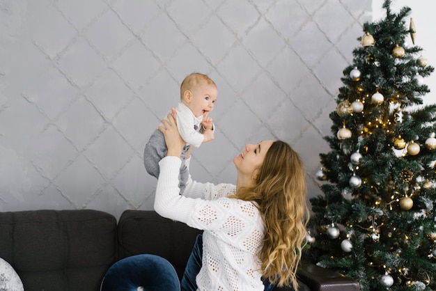La jeune mère a élevé son fils dans ses bras et lui sourit. Ils célèbrent Noël et le nouvel an dans le salon avec un arbre de Noël