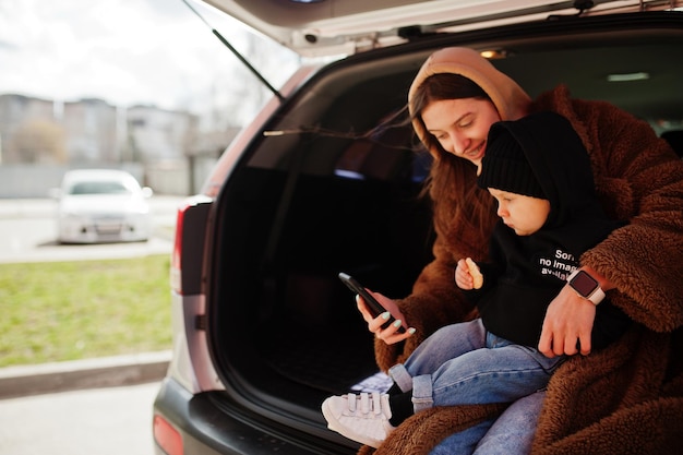 Jeune mère et enfant assis dans le coffre d'une voiture et regardant un téléphone portable Concept de conduite de sécurité