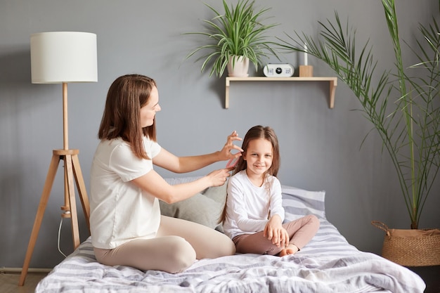 Jeune mère faisant la coiffure à sa fille assise sur le lit dans la chambre à peigner les cheveux exprimant des émotions positives se préparant à aller à l'école