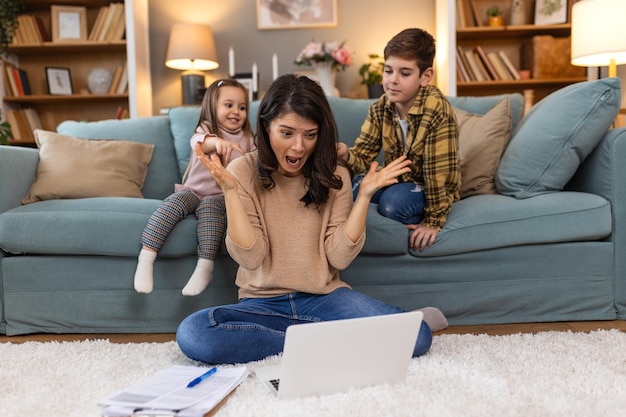 Une jeune mère fatiguée assise sur le sol et travaillant avec un ordinateur portable et des documents pendant que les petits enfants sautent sur le canapé et s'amusent et font du bruit