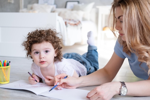 Jeune mère et fille enfant bouclée dessinant avec des marqueurs de couleur allongés sur le sol à la maison