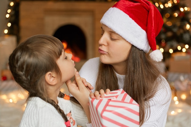 Jeune mère et fille séduisantes avec boîte-cadeau se regardant, veut embrasser sa mignonne petite fille, la veille de Noël à la maison, posant près de la cheminée.