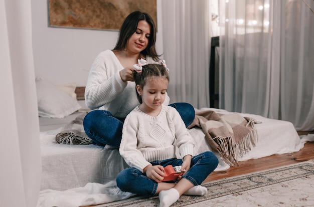 Une jeune mère heureuse fait la coiffure de sa petite fille assise sur le lit.