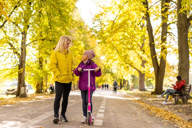 Jeune mère marchant avec sa fille sur un scooter pour tout-petits dans le parc.