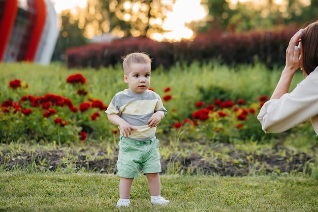 Une jeune mère mignonne aide et apprend à son petit fils à faire ses premiers pas pendant le coucher du soleil dans le parc sur l'herbe.