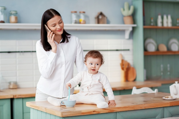 Jeune mère, parler au téléphone à la maison dans la cuisine en regardant son bébé