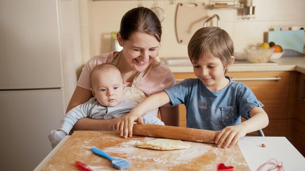 Photo jeune mère avec un petit bébé regardant son fils aîné rouler la pâte et cuisiner