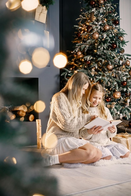 Jeune mère et petite fille assise sur un sapin de noël à la maison et lisant un livre