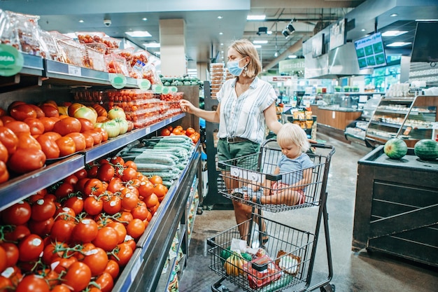 Une jeune mère portant un masque de protection achète de la nourriture avec un bébé dans le panier d'achat
