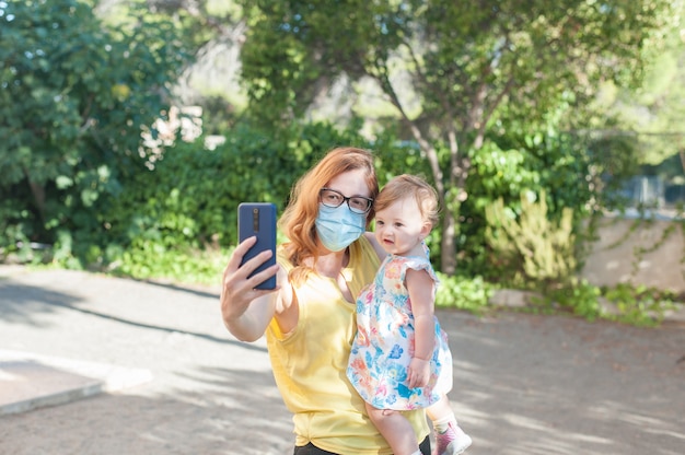 Jeune mère portant un masque de protection avec son bébé dans ses bras à l'aide d'un téléphone portable à l'extérieur