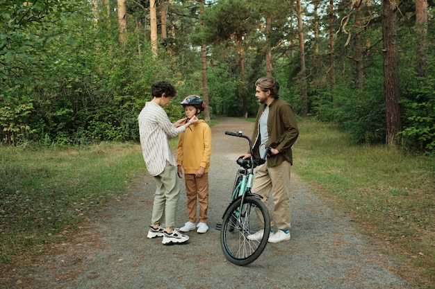 Jeune mère prudente aidant son fils à attacher les ceintures de casque de sport pendant que son père se tenait à vélo sur la route entre les arbres verts