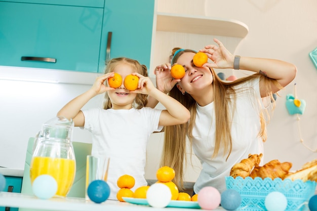 Une jeune mère et sa fille dans la cuisine à la table jouent avec des mandarines d'agrumes frais au lieu des yeux sur leurs visages Relation familiale avec un enfant