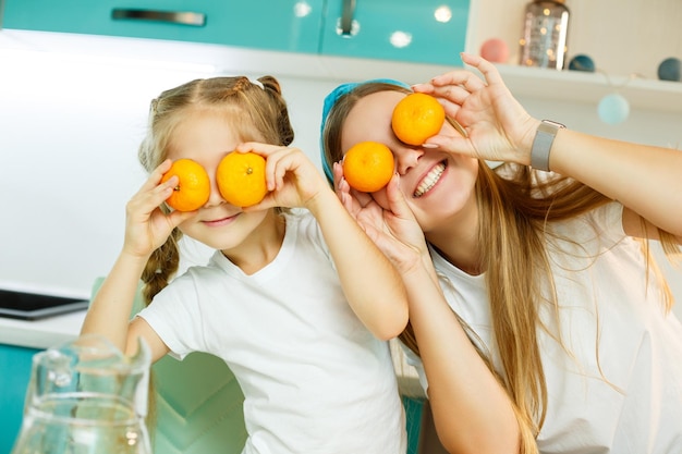 Une jeune mère et sa fille dans la cuisine à la table jouent avec des mandarines d'agrumes frais au lieu des yeux sur leurs visages Relation familiale avec un enfant