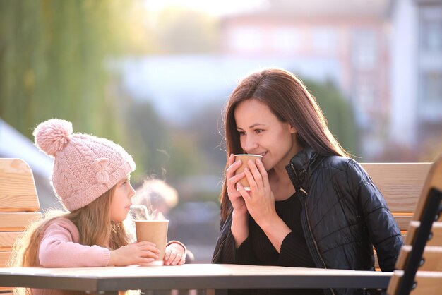 Jeune mère et sa fille enfant s'amusant ensemble assis au café de la rue avec des boissons chaudes le jour d'automne ensoleillé Bonheur dans le concept des relations familiales