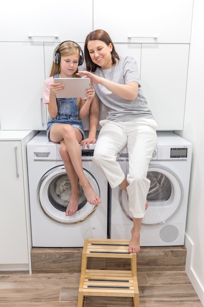 Jeune mère et sa fille jouent avec une tablette dans la buanderie assis sur une machine à laver