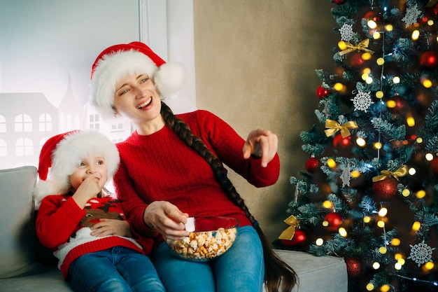 Une jeune mère et sa fille portant des chapeaux de Père Noël regardent des films à la maison pour Noël et mangent du pop-corn