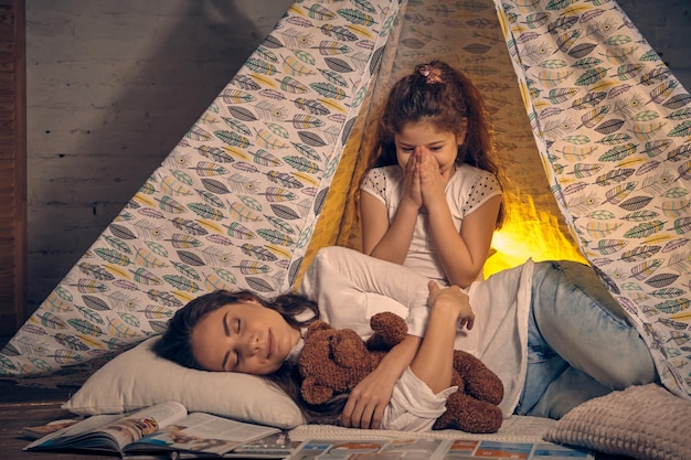 La jeune mère et sa jolie fille sont dans une tente tipi avec des oreillers. Maman dort avec un ours en peluche. La fille sourit et la regarde. Famille heureuse.