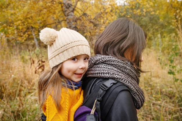 Jeune mère avec sa petite fille sur le dos dans un porte-bébé ergonomique dans la nature d'automne Portage et concept de mère active