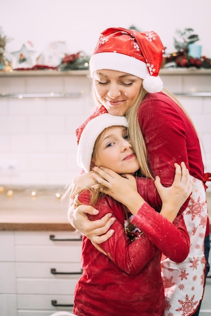 Une jeune mère se tient avec son enfant adolescent en chandails rouges dans la cuisine étreignant en chapeaux de père Noël