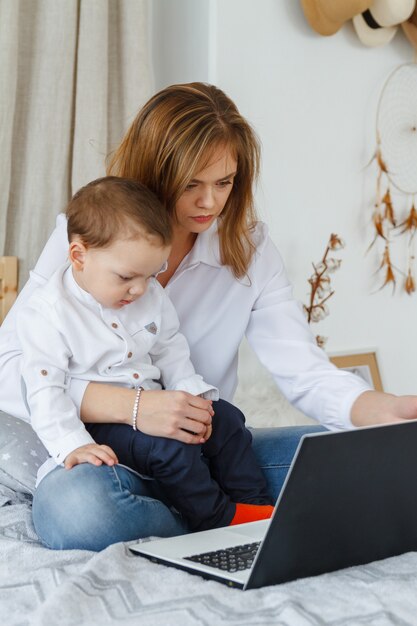 Une jeune mère avec son fils bien-aimé dans la chambre sur le lit avec un ordinateur portable. Travailler à la maison avec un concept d'enfant