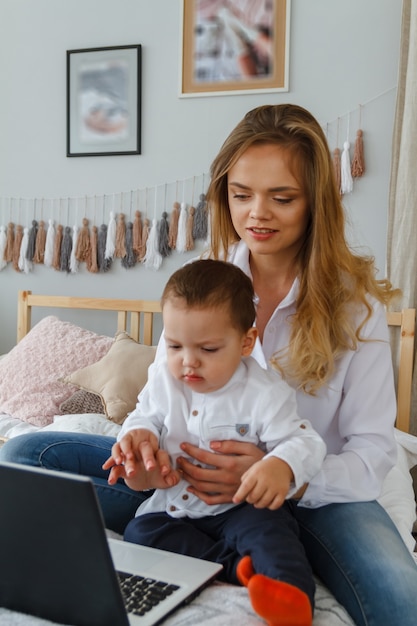 Une jeune mère avec son fils bien-aimé dans la chambre sur le lit avec un ordinateur portable.