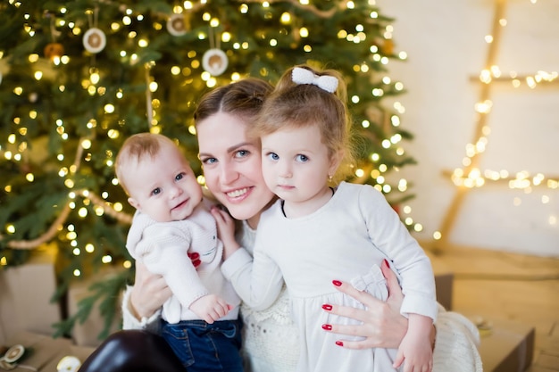 Jeune mère avec son fils et sa fille près de l'arbre de Noël Héhé en attendant Noël et le Nouvel An Intérieur lumineux confortable avec des décorations et des lumières de Noël