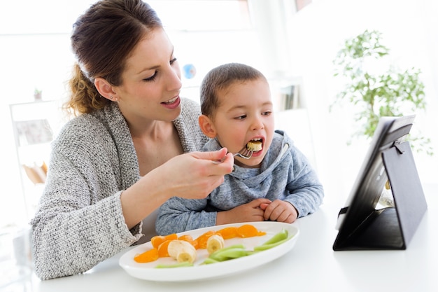 La jeune mère et son fils utilisent une tablette numérique tout en mangeant des fruits à la maison.