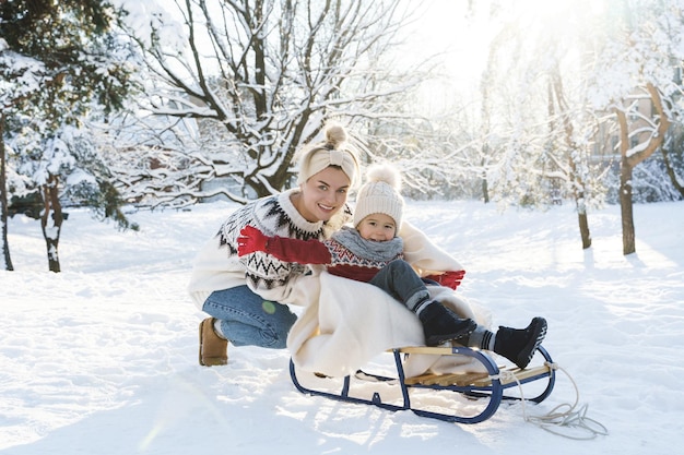 Jeune mère et son mignon petit fils avec traîneau rétro dans un parc enneigé pendant la journée d'hiver ensoleillée