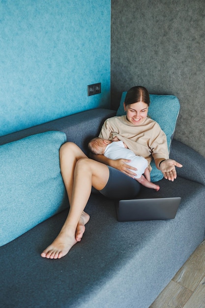 Une jeune mère souriante allaite son bébé et travaille à la maison avec son bébé sur le canapé tout en écrivant un e-mail allaitant le bébé Une jeune femme travaille à la famille